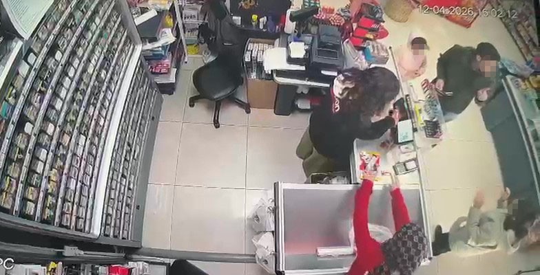 Bir marketteki kasa alanında, çalışan bir kadın müşteriye yardımcı oluyor. Müşteriler, kasanın önünde çeşitli ürünlerle etkileşimde bulunuyor. Arka planda market rafları görünmekte ve birçok ürün var.