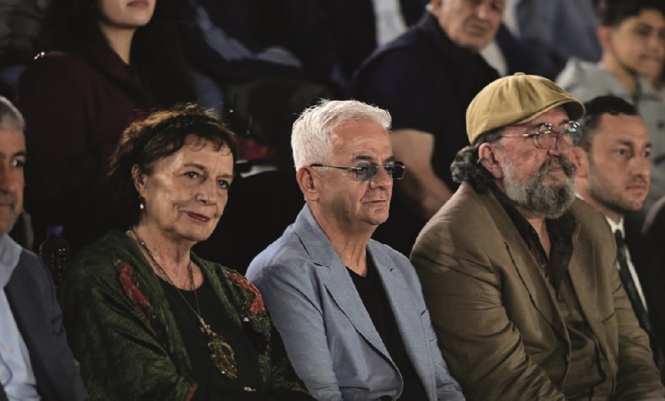Bir grup insan, bir etkinlikte oturuyor. Ön planda üç kişi, erkek ve kadın, ilgiyle bir şeyleri izliyorlar. Arka planda, kalabalık bir seyirci görünmekte.