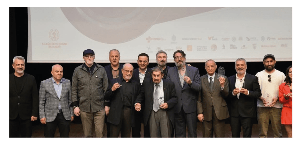 Bir ödül töreninden görüntü, sahnede çeşitli bireyler ödüllerini tutarak poz veriyor.