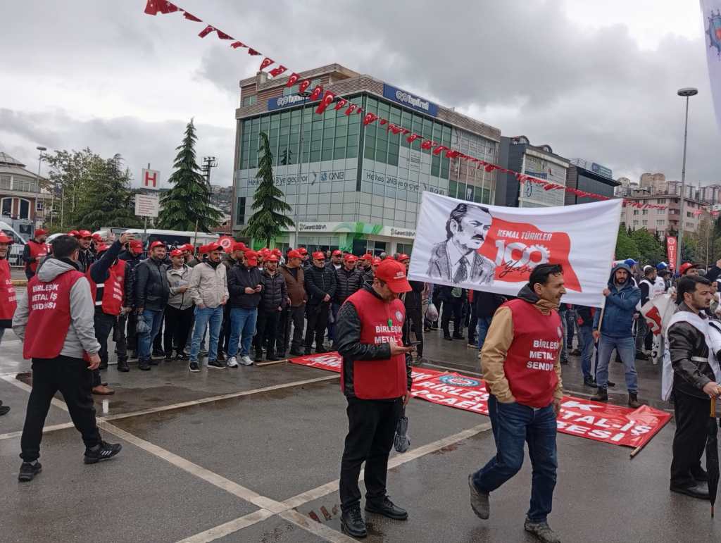 Bir grup insan, kırmızı şapka ve yelek giymiş, bir protesto etkinliği düzenlemektedir. Arkada bir pankartta Atatürk'ün resmi yer almakta. Etkinlik alanında Türk bayrakları dalgalanmakta.