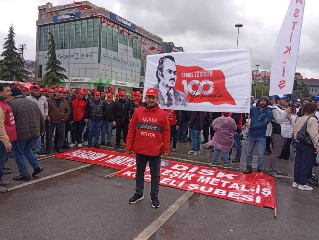 Bir grup insan, Kemal Türker'in 100. doğum yılı etkinliğinde toplanmış. Katılımcılar kırmızı şapkalar ve tişörtler giymekte. Bir pankart ve dikkat çekici bir afiş ile beraber kalabalık bir miting atmosferi var.