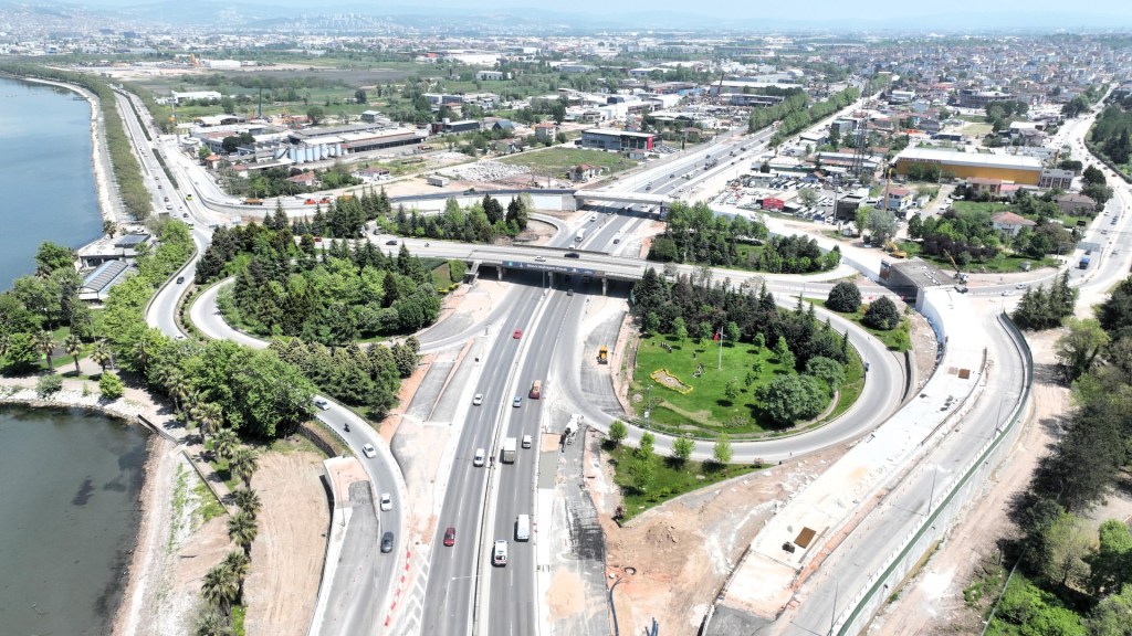 Hava fotoğrafı; bir kavşak, yollar, yeşil alanlar ve bir nehir görünmektedir. Şehir manzarası arka planda yer almaktadır.