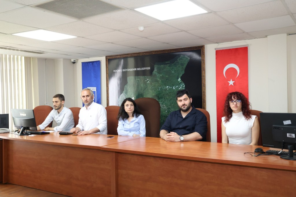 Beş kişi, Kocaeli Büyükşehir Belediyesi'nde bir toplantıda oturuyor. Arkada Türkiye bayrağı ve belediyenin haritası yer alıyor.