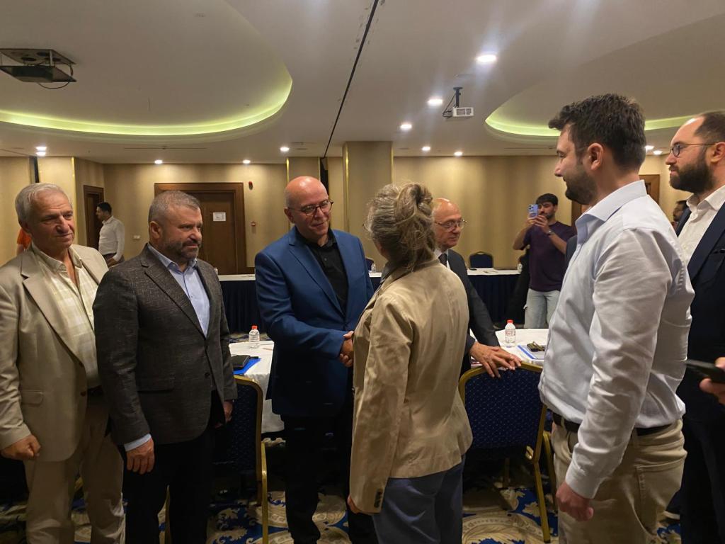 Bir grup insan, bir toplantı odasında selamlaşıyor ve sohbet ediyor.